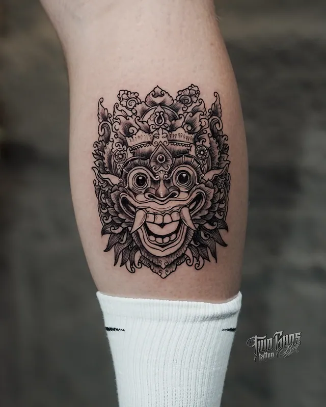 Black and grey ornamental Barong mask calf tattoo framed by scrollwork and lotus petals