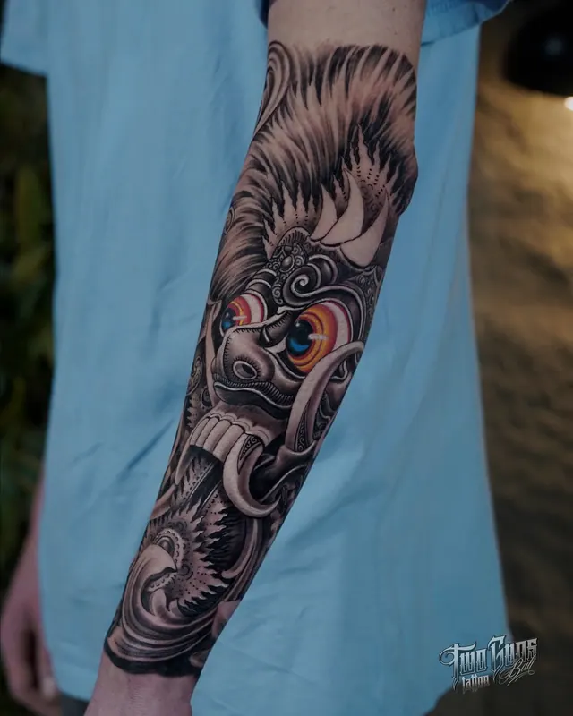Balinese Rangda mask forearm sleeve tattoo in black and grey with colour eye accents and fangs