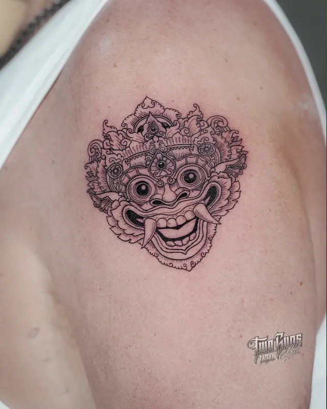 Fine line Balinese Barong mask shoulder tattoo with fangs, a crown and ornamental scrollwork