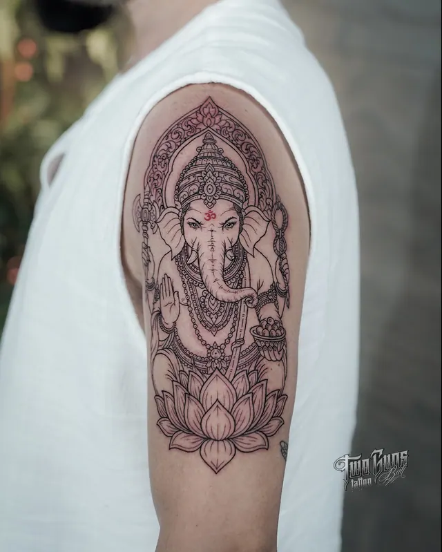 Fine line Ganesha upper arm tattoo seated on a lotus, with ornamental detail and red accents