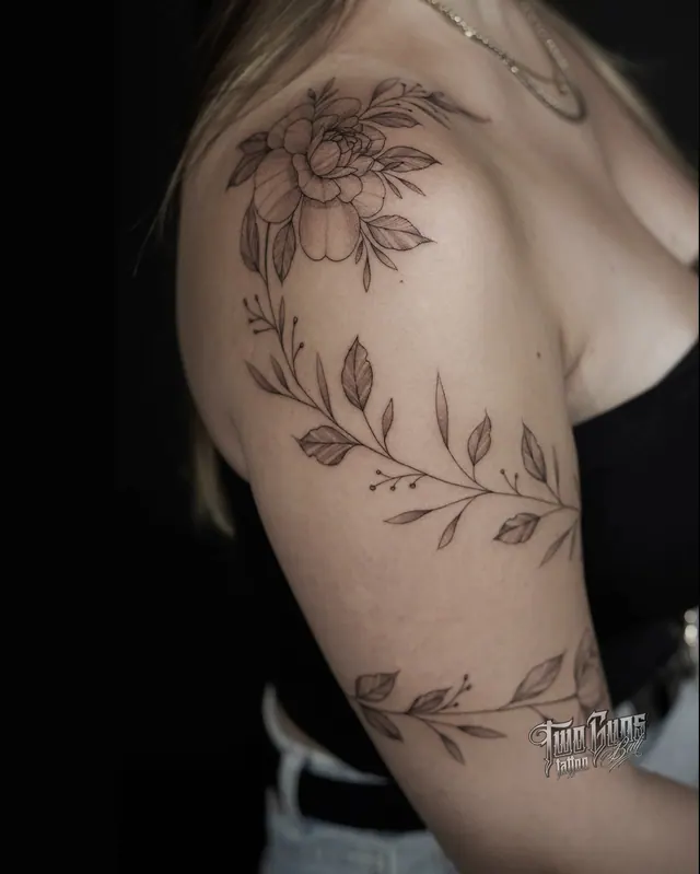 Fine line floral tattoo on a woman's shoulder and upper arm, a peony with trailing leaf vines