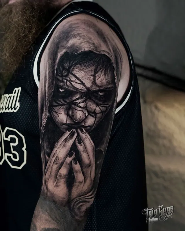 Black and grey horror tattoo of a hooded faceless figure with clasped hands, on the upper arm